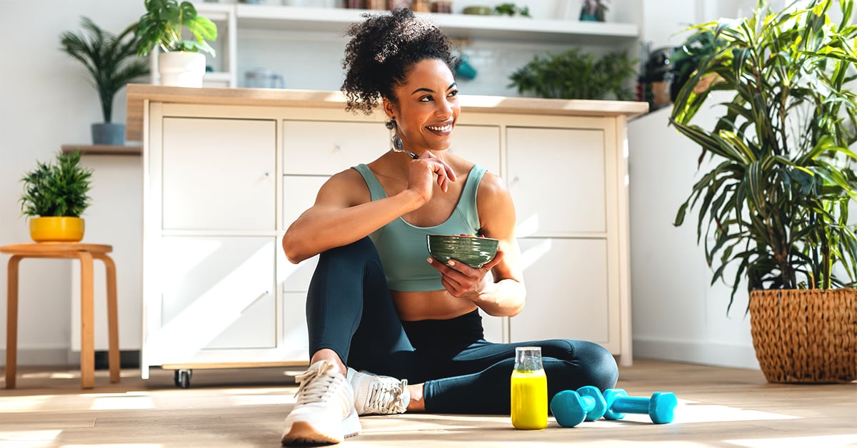 A woman sitting on the floor eating a bowl of fruit with a bottle of juice and two dumbbells sitting in front of her.