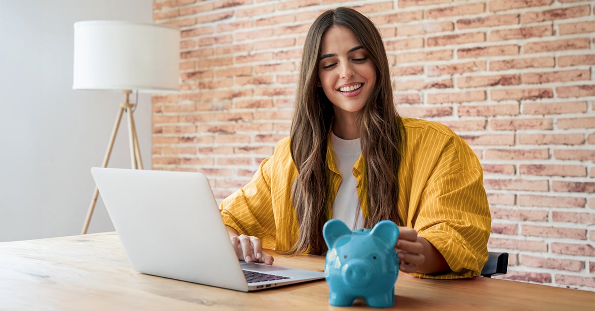 A woman sitting at a desk with a laptop and piggy bank.