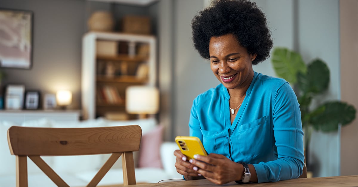 A woman sitting at a table looking down at a smartphone, smiling.