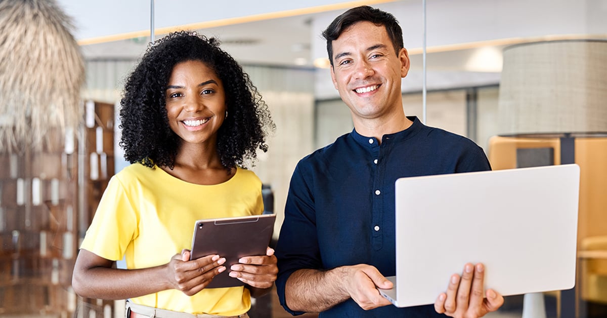 A man and a woman standing and smiling, holding a tablet and laptop.
