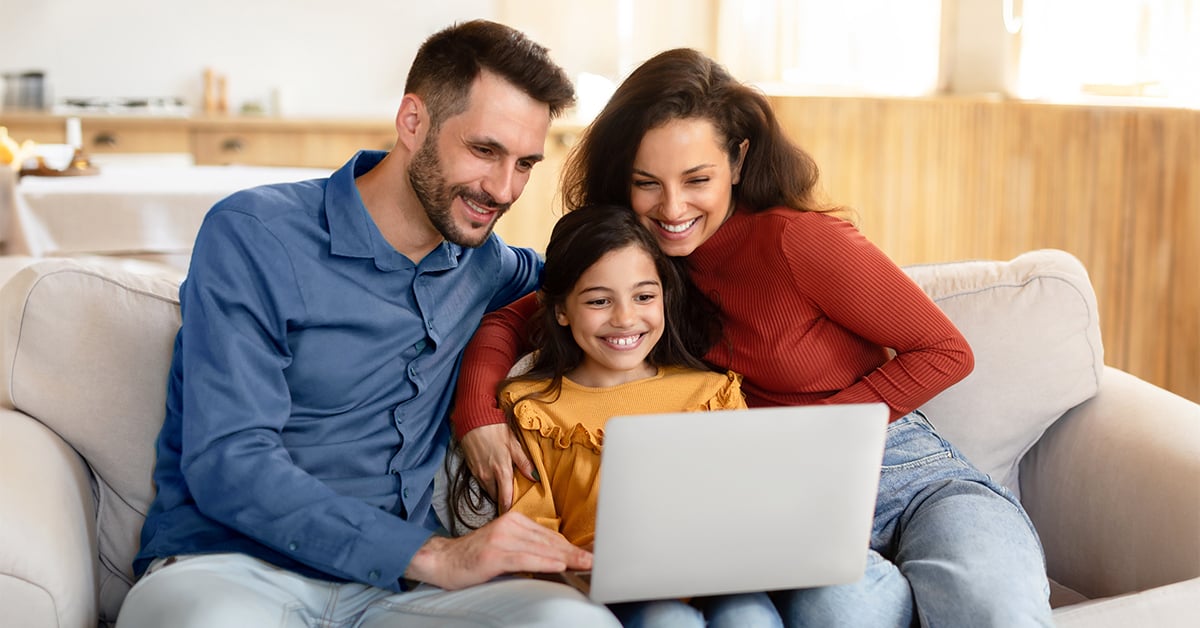 A family of three sitting on a couch in front of a laptop computer, smiling.